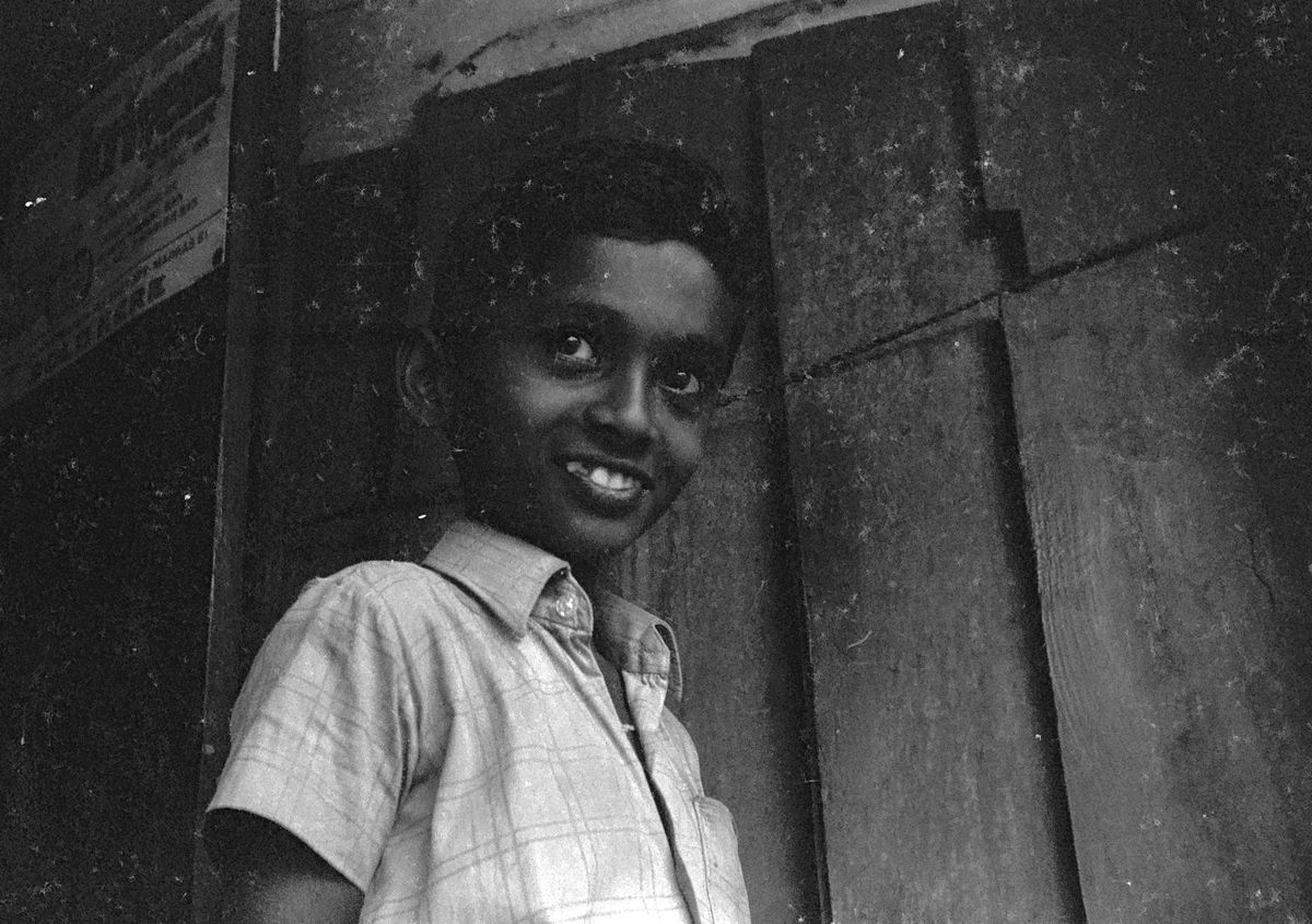 A boy in a checked shirt smiling shyly from a dark wooden doorway.