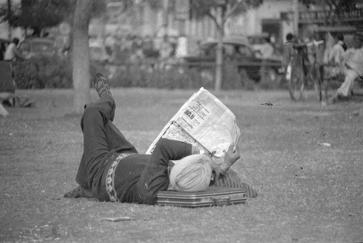 A man lying on the grass in a park, his head on a bag, reading a newspaper held up over his face.