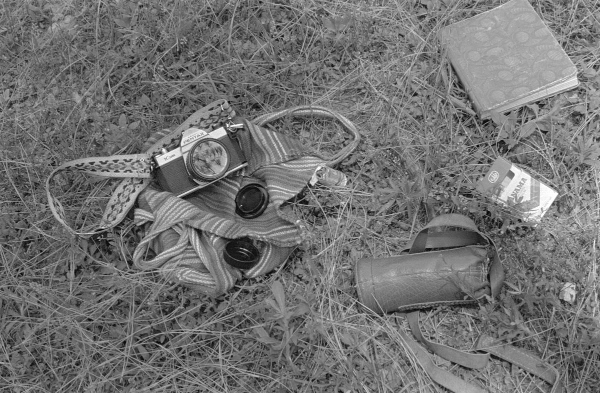 An old film camera lying in the grass with its strap, a lens cap and a notebook beside it.
